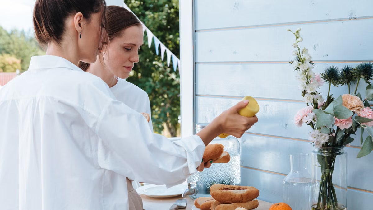 Women in white outfits preparing a meal outdoors with breads and citrus fruits. Bright and lively setting.